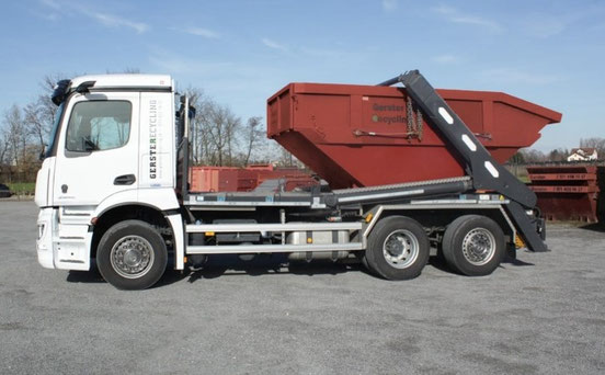 Ein weisser Lastwagen von Gerster Recycling mit Mulde, Container, Box. Im Hintergrund sind mehrere grosse rote Abfallcontainer mit dem Logo von Gerster Recycling zu sehen. Das Bild zeigt einen Recycling- und Abfallentsorgungsbetrieb.