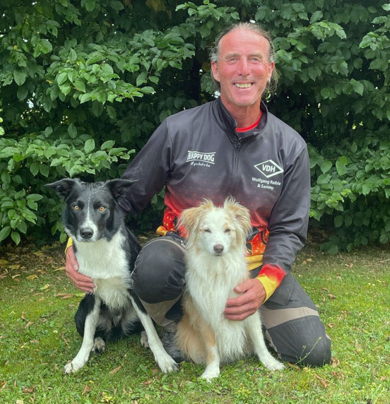 Wolfgang kniet auf Gras vor einem Busch und hält seinen schwarz-weißen Hund Boh und seinen hellbraun-weißen Hund Sammy an den Seiten fest.