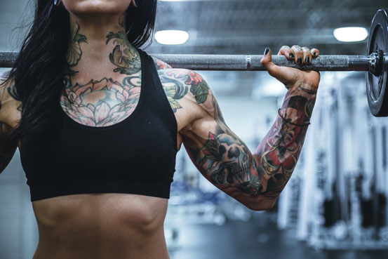 Der Oberkörper einer Frau im Fitnessstudio. Auf den Schultern hält sie eine Langhantelstange und trainiert. Sie trägt ein schwarzes und bauchfreies Top. Ihr Oberkörper ist tätowiert.