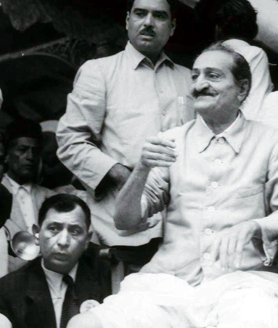 Minoo Kharas sitting next to Meher Baba with Eruch Jessawala standing behind assisting.
