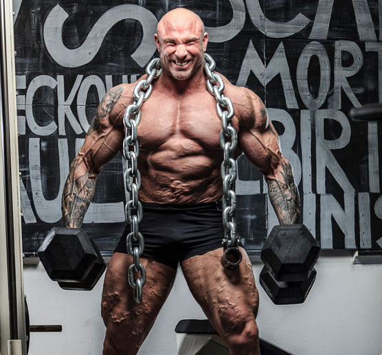 Ein Bodybuilder hält zwei Kurzhantel in der rechten und linken Hand. Um den Hals hängt eine schwere Eisenkette. Bis auf eine schwarze Shorts trägt er nichts. Seine Muskeln sind sehr deutlich zu sehen.