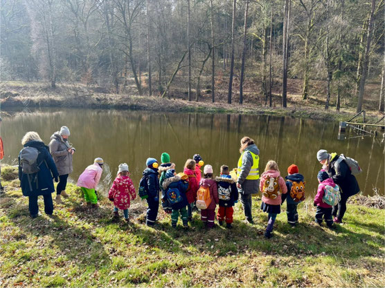 Ein Dutzend Kinder, von hinten fotografiert, steht am Rand eines sonnenbeschienenen Teichs