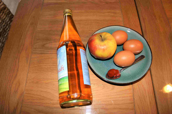 Auf einem Holztisch liegt ein blauer Teller, auf dem ein Apfel, drei Eier und ein Teelöffel mit Tomatenmark liegen. Neben dem Teller liegt links eine Flasche Apfelsaft.
