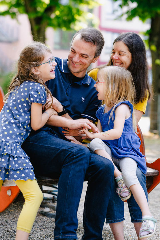 Eine vierköpfige Familie sitzt auf einem Spielplatz beisammen. Sie kitzeln sich gegenseitig, dabei müssen alle herzlich lachen.