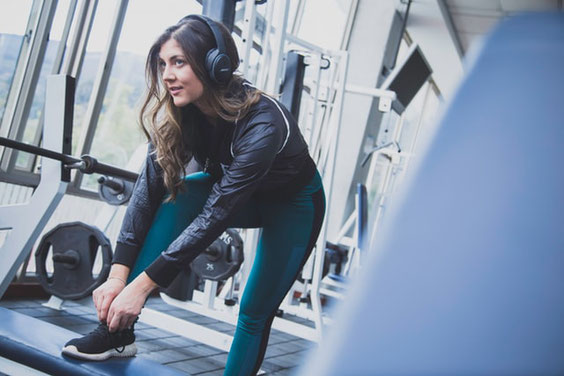 Eine Frau im Fitnesstudio. Sie hat ihren rechten Fuß auf einer Kiste aufgestellt, um sich den Schuh zuzubinden. Sie bereitet sich auf das Training vor und trägt Kopfhörer, um Musik zu hören.