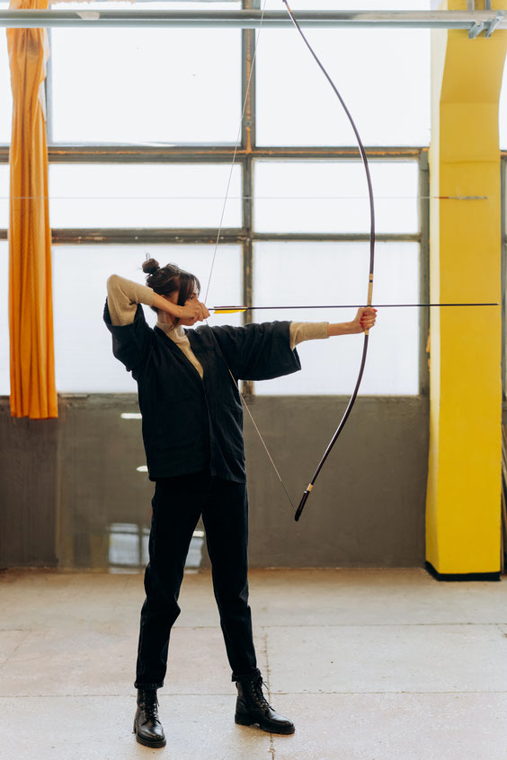 A woman practices archery indoors, drawing a traditional longbow with precision. She wears a dark kimono-style jacket, black pants, and boots.