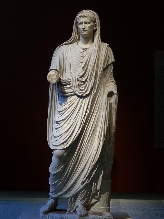 Marble statue of a Roman man in a draped toga with a covered head, missing both hands, standing against a dark background.