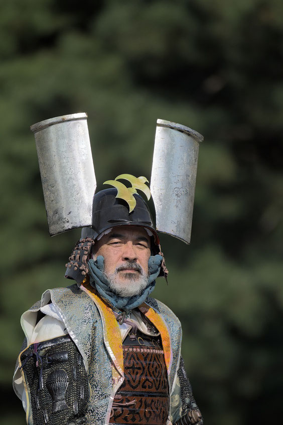 A samurai warrior in elaborate armor wears a striking helmet adorned with two large cylindrical ornaments and a golden emblem.
