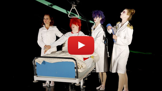 Theaterbühne: 3 Ärztinnen um einen im Krankenbett sitzenden Patienten