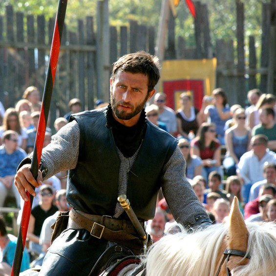 A man in medieval attire holding a lance rides a horse during a jousting event, with a crowd watching in the background.