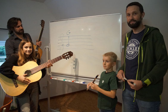 Zwei Kinder und zwei Erwachsene mit Gitarren und Querflöten vor einer Tafel