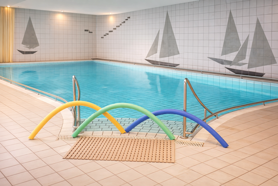 Innen-Schwimmbecken mit blauen Wasser, umgeben von weißen Fliesen, bunte Schwimmnudeln in Gelb, Grün und Blau liegen am Beckenrand, Wand mit Segelboot-Motiven