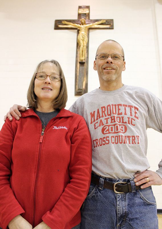 Chris and Kathie Lampe pose for a photo with the recently blessed, handcrafted crucifix now located in the gymnasium of the Marquette Education Center.