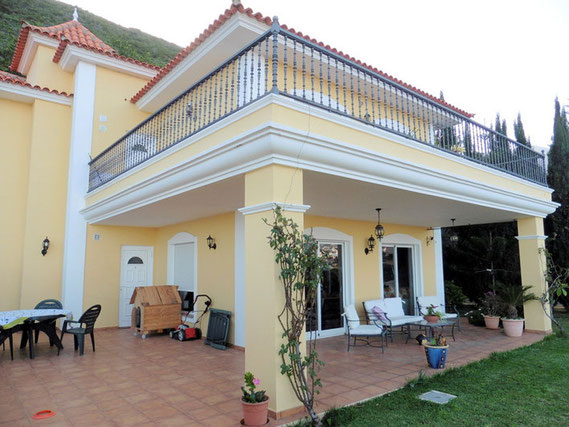 Blick vom Garten auf das 2 Stöckige Haus mit Terrasse und darüber einem Balkon. Die Immobilie ist im gelben Farbton gestrichen und die Fenster sind weiß eingefasst.