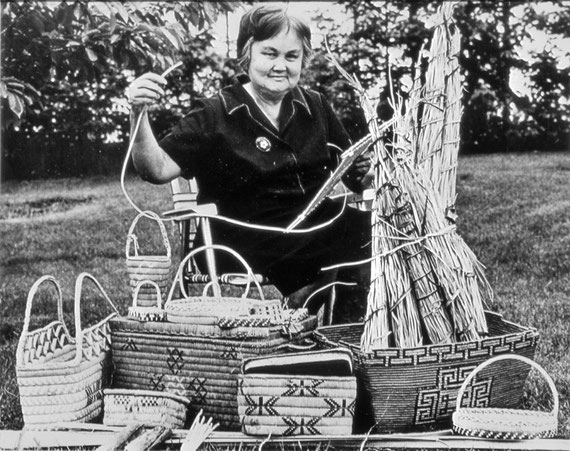 This photo from the 1970s shows Eva May Nahanee with examples of her basketry work and bundles of raw cedar roots.