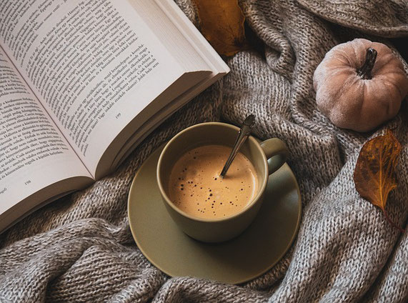 Ein herbstliches Foto. Auf einer Strickdecke liegt ein aufgeschlagenes Buch, ein kleiner brauner Kürbis und Laub. Daneben steht eine Tasse Kaffe.