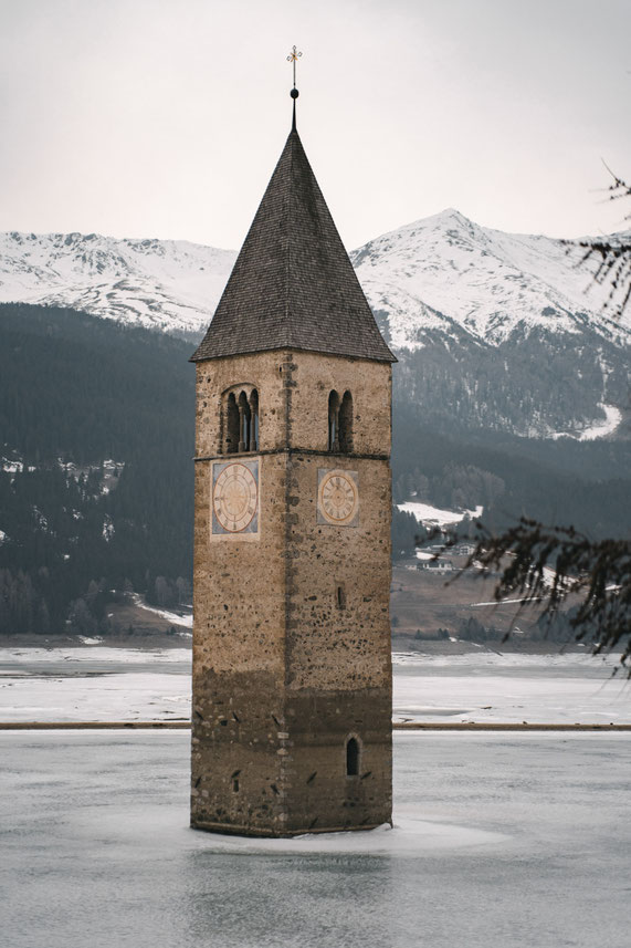 An old church tower rises from a frozen lake against a backdrop of snow-capped mountains.