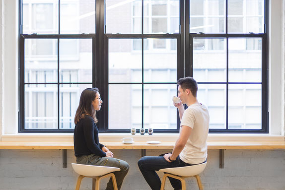 Un homme et une femme sont assis dans un endroit lumineux. La jeune femme parle vers le jeune homme qui boit un café.