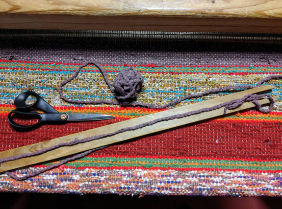 Tapis en lirette à rayures rouges, verts et chinés, en train d'être tissé sur un métier à tisser. Sur la surface déjà tissée, sont posées une grande navette en bois, une pelote de bande de tissu violet et la paire de ciseaux.