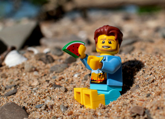 männliche Legofigur sitzt am Strand, Melonenstück in Hand
