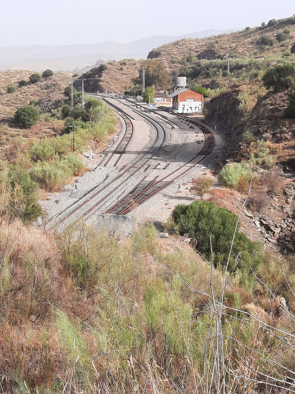 Das sind 2 Bahnhöfe: die im Nichts endenden Gleise, der Wassertank und ein verfallendes Gebäude gehören zur Dampflokzeit. Und das neue Gleis und das größere Gebäude in unsere Zeit.  Ich weiß nur nicht,  woher die Passagiere kommen sollen. Kein Ort weit und breit.  Aber das war in Namibia genauso  😉. Die Gleise verschwinden unter mir im Tunnel. 