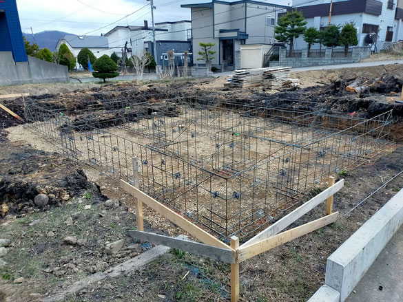 住宅が並ぶ背景を背に、反対側から斜め構図で撮影された基礎配筋全体。奥には積み重ねられた板も見える