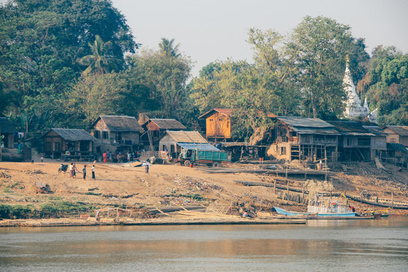 Auf dem Foto ist ein Dorf in Myanmar zu sehen. Es sind nur wenige Hütten am Flußufer. Dahinter befindet sich ein Wald.