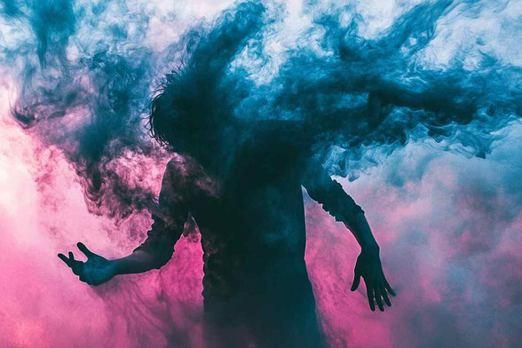 Eine Person mit ausgestreckten Armen, um die herum Rauch in Blau- und Rosatönen wirbelt und eine jenseitige Atmosphäre schafft