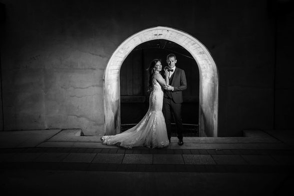 Black and white photo of a bride and groom in front of a lit archway. Bride is in a white dress and groom is in a suit. 