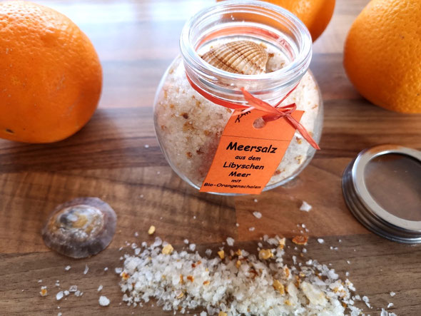 Meersalz mit Orangenschalen im Glas mit Muschel und orangem Etikett von 2 Orangen flankiert