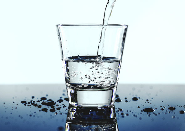 Ein Glas Wasser, das befüllt wird. Es steht auf einem blauen Tisch mit spiegelnder Oberfläche.