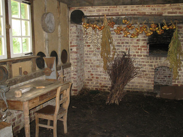 Binnenzicht oude hoeve. Met een tafel en één stoel. Een bussel hout en gedroogde ajuin boven de oven.