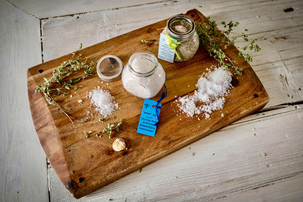 Meersalz und Kräutersalz im Glas und auf einem Holzbrett verstreut, mit blauem Etikett und Oregano verziert