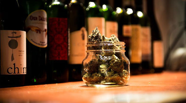 Ein Glaß steht gefüllt mit Cannabis auf einem Holztresen. Im hintergrund befinden sich Weinflaschen