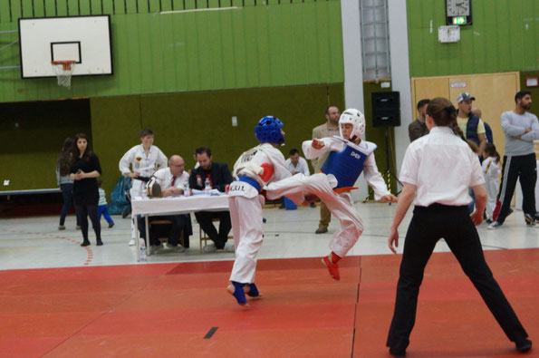 Zwei Kinder kämpfen auf einem Taekwondo-Turnier. Der Junge mit dem weißen Helm macht einen Sprungtritt gegen den Jungen mit dem blauen Helm.