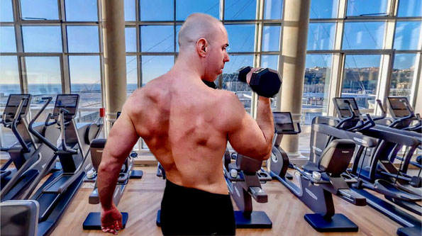  Un athlète musclé réalisant des curls avec haltères dans une salle de sport de prestige avec vue panoramique sur la mer à Monaco, illustrant un entraînement de force d’élite et un coaching personnalisé.
