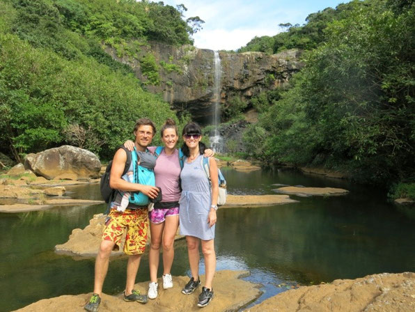 Glückliche Familie mit Tragepapa vor Wasserfall während der Elternzeit-Reise.