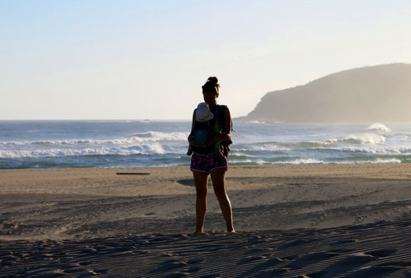 Glückliche Mutter mit Baby in der Trage am Strand während ihrer Elternzeit-Reise.