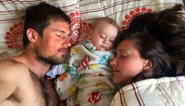 Junges Paar schläft mit gemeinsamem Baby in der Mitte im Bett während Elternzeit-Reise.