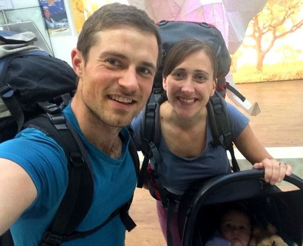 Junge Familie unternimmt während der Elternzeit die erste Reise mit Baby & lächelt am Flughafen in die Kamera.