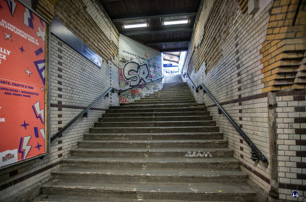 Blick nach oben zum Bahnsteig. Die originären weiß oder braun glasierten Ziegelsteine sind größtenteils noch erhalten, wenn auch im oberen Bereich lückenhaft. Auch die Handläufe zu beiden Seiten der Treppe mit ihren kunstvollen Befestigungselementen sind 