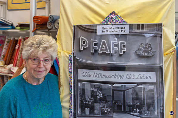 Die heutige Inhaberin, Frau Monika Tschernoch, vor einem Foto, welches das Ladengeschäft im Eröffnungsjahr 1955 zeigt. Sie selbst hat den Laden von der Fa. Pfaff 1996 übernommen.