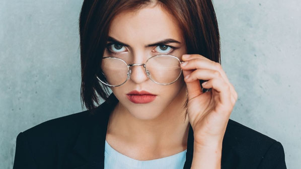 Eine Frau mit kurzen braunen Haaren und intensiven Augen blickt direkt in die Kamera und zieht ihre Brille leicht nach unten. Ihr ernster, fragender Ausdruck symbolisiert Selbstreflexion und die Auseinandersetzung mit der eigenen Identität jenseits der Fü