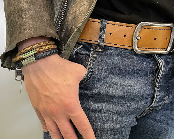 Am rechen Handgelenk eines Mannes befindet sich ein einfach gewickeltes schwarzes Lederarmband mit Edelstahl Magnetverschluss und darüber ein goldfarbenes doppeltes Lederarmband mit Edelstahl Magnetverschluss 
