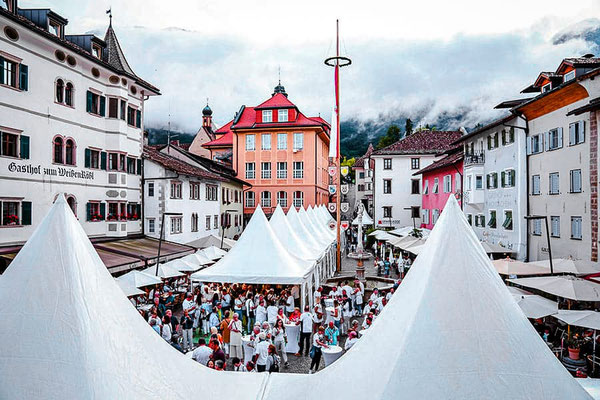 Der Marktplatz in Kaltern während der Veranstaltung „Kaltern ganz in Weiß“, mit festlich aufgebauten Zelten und einer großen Menschenmenge, die die besten Kalterer Weißweine verkostet und genießt. Im Hintergrund sind die historischen Gebäude von Kaltern z