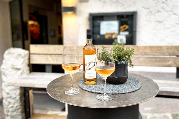 Vino rosato della cantina Passir su un tavolo nella Val Passiria, due bicchieri pieni accanto alla bottiglia e a un vaso di erbe aromatiche, un'elegante scena di piacere culinario all'aperto