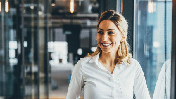 Eine lächelnde, selbstbewusste Frau in einem weißen Hemd steht in einem modernen Büro mit Glaswänden. Die offene und freundliche Ausstrahlung symbolisiert moderne Führungskompetenzen, Selbstbewusstsein und eine positive Arbeitskultur