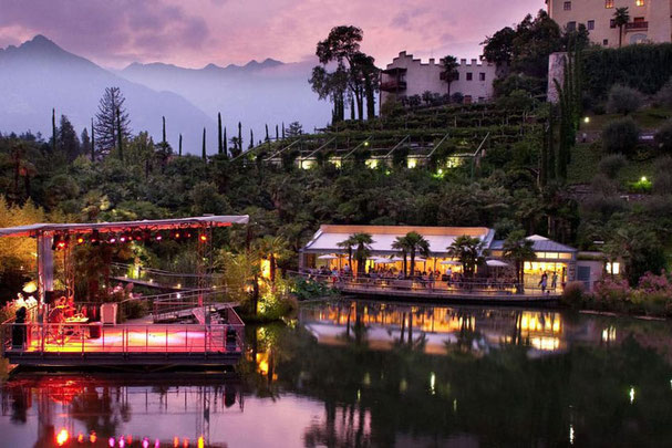 Atmosfera serale nei giardini di Trauttmansdorff con palco, lago e ristorante illuminato sullo sfondo delle montagne.