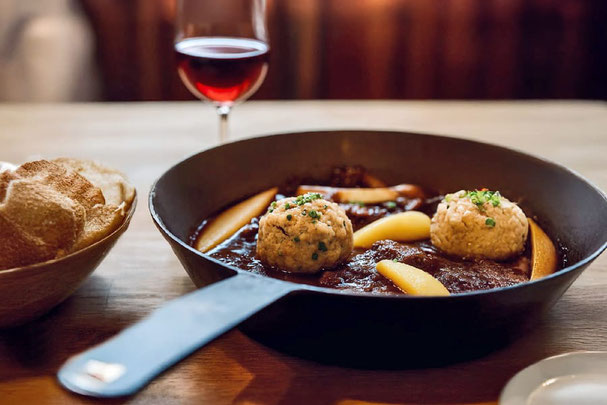 Schmorgericht vom Schnalser Schaf mit Knödeln und Birnen in Pfanne, serviert mit Brot und Rotwein auf rustikalem Holztisch.