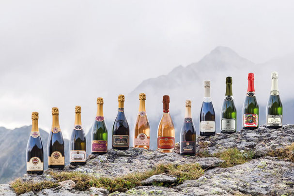 Una serie di bottiglie della cantina di spumanti Arunda, che mostrano diverse annate e varietà di spumante altoatesino, su una roccia davanti a un imponente scenario montano.